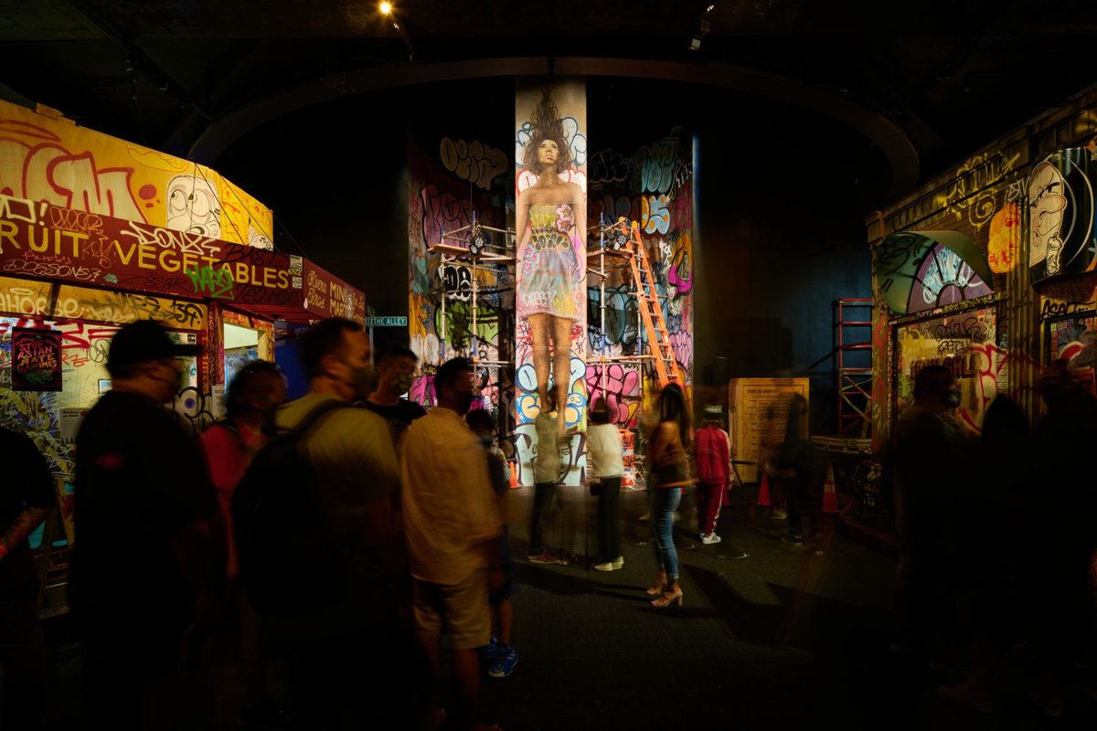 Visitors walk through a dim indoor streetscape lined with painted storefronts, murals and a mural-covered column.