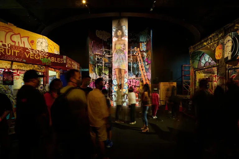 Visitors walk through a dim indoor streetscape lined with painted storefronts, murals and a mural-covered column.